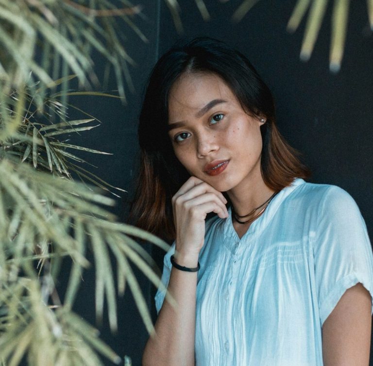 Lady in white. Young woman in a light blue blouse poses thoughtfully beside greenery.