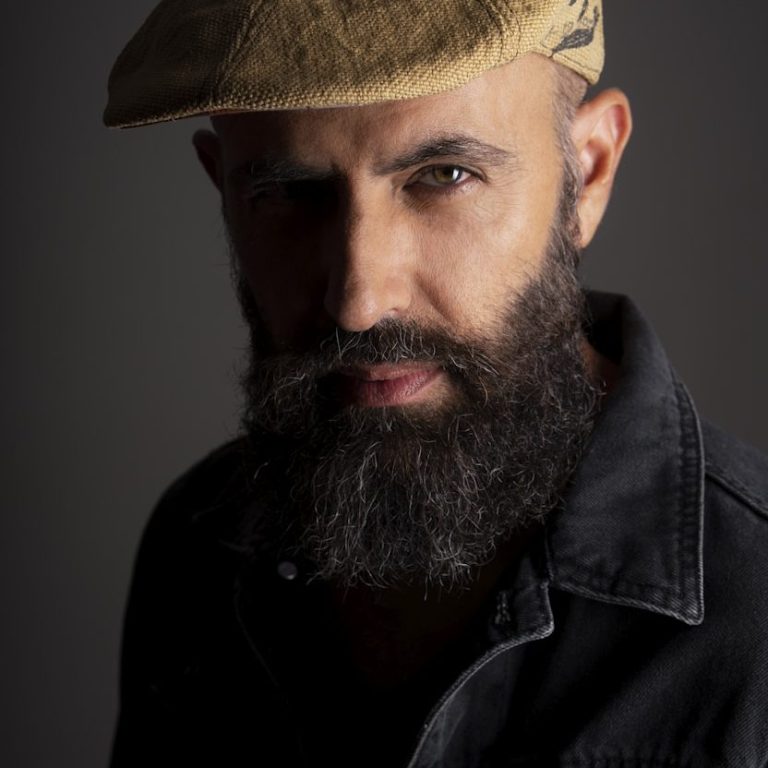 Bearded Man in Peak Cap Man with a beard and a cap, wearing a black shirt, against a dark background.